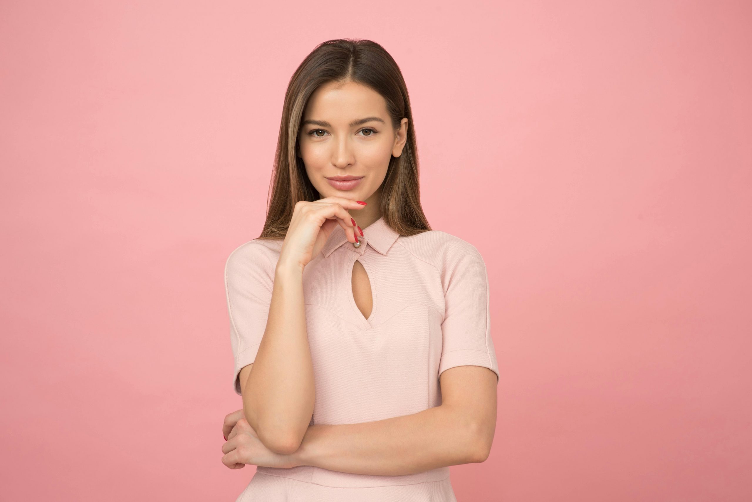 woman in a blush pink dress smiling charmingly, with one hand elegantly placed on her chin